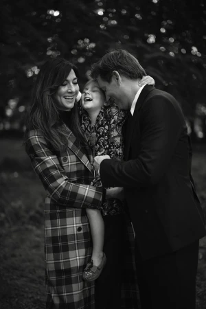 Black and white portrait of a smiling family of three hugging outdoors in a park with trees in the background.