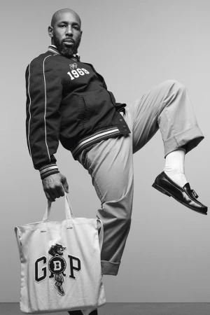 Black man with a beard wearing a letterman jacket and holding a GAP tote bag while lifting his leg in a studio setting.