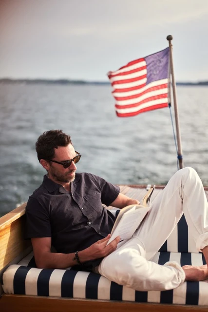 Adult male reading a book on a wooden boat with an American flag, wearing a dark shirt, white pants and sunglasses in daylight.