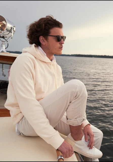 Young man in sunglasses, wearing a hoodie and pants, sits on a boat against a water and sky background, creating a relaxed outdoor scene.