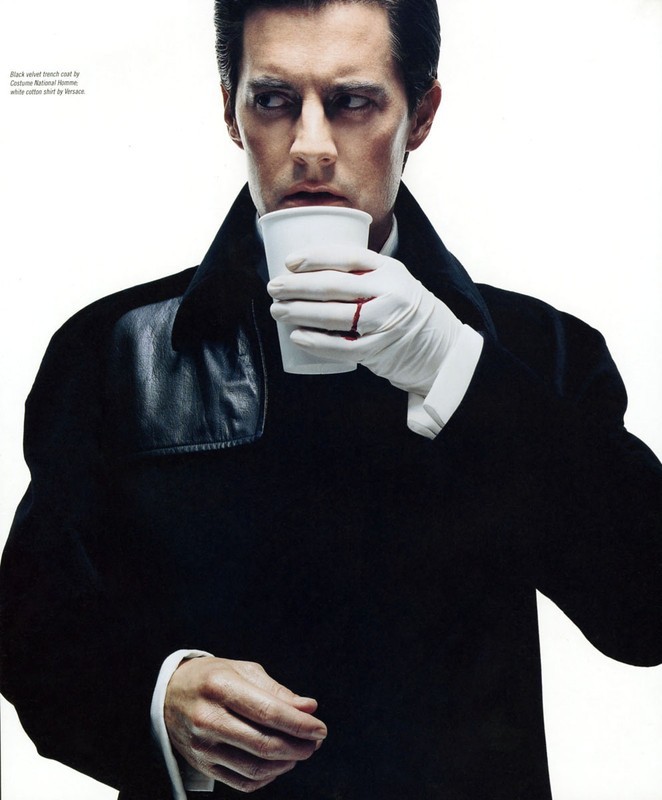 Fashion shot of a man in a black coat, white shirt and gloves, drinking from a white cup against a stark white background.