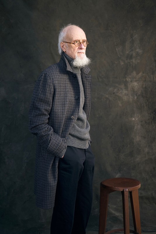 Portrait of an adult man in a patterned overcoat and eyeglasses, standing near a stool against a mottled grey studio background.