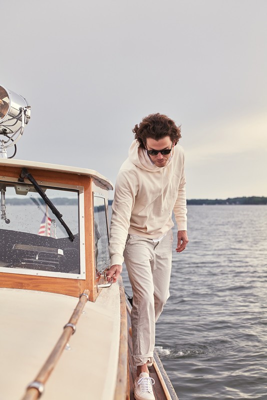 Man on a boat wearing a light-colored hoodie and pants walks on the deck near the cabin, with the sea visible in the background.
