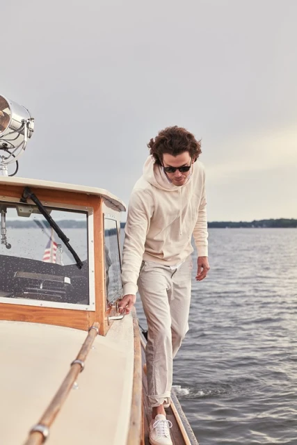 Man on a boat wearing a light-colored hoodie and pants walks on the deck near the cabin, with the sea visible in the background.