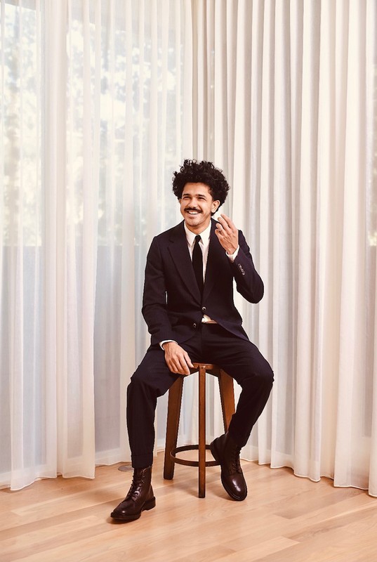 A man in a dark suit, tie, and lace-up boots sits on a stool against a neutral curtain backdrop, captured in a medium studio shot.