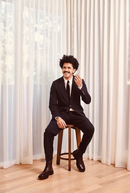 A man in a dark suit, tie, and lace-up boots sits on a stool against a neutral curtain backdrop, captured in a medium studio shot.