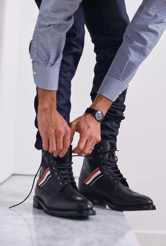 Close-up of a man tying the laces of stylish black leather boots featuring red, white, and blue stripes, worn with dark jeans and a button-down shirt.