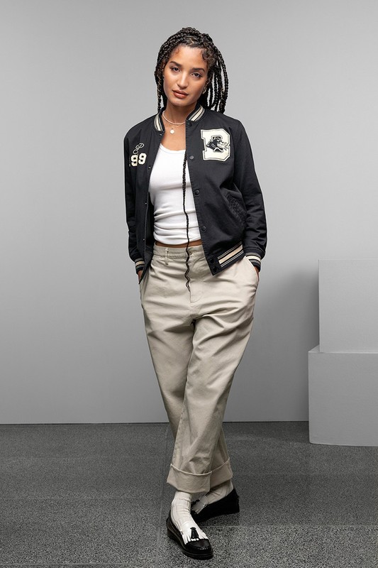 Full-length studio shot of a young woman with braided hair posing in a black jacket, white top, and khaki pants against a gray backdrop.