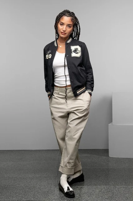 Full-length studio shot of a young woman with braided hair posing in a black jacket, white top, and khaki pants against a gray backdrop.