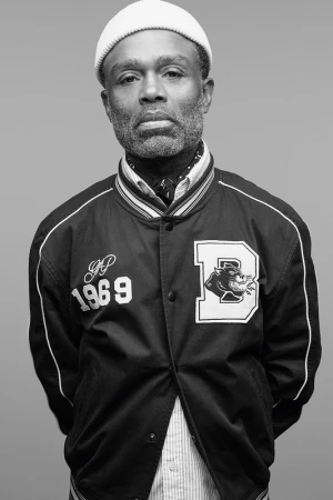 Black man wearing a beanie and a letterman jacket against a plain grey backdrop in Los Angeles.