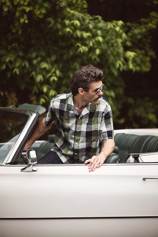 Man with a mustache and sunglasses exits a white convertible car with green interior and trees in the background on an overcast day.