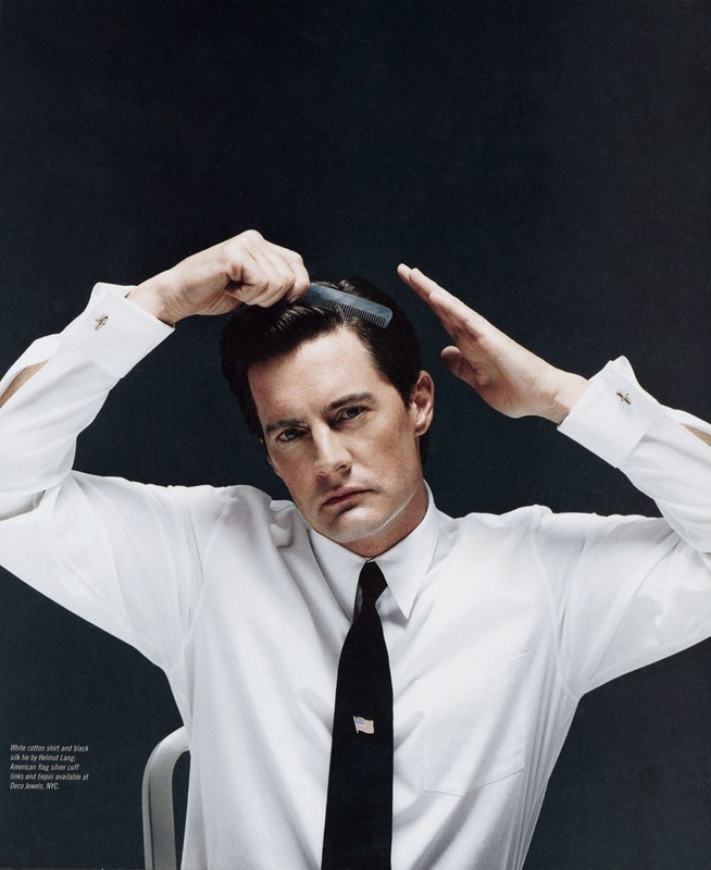 Close-up portrait of a man in a white shirt and black tie, combing his hair with a black comb against a dark grey background.