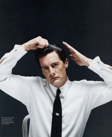 Close-up portrait of a man in a white shirt and black tie, combing his hair with a black comb against a dark grey background.