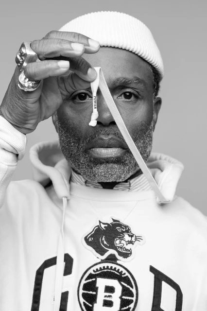 Monochrome portrait of a Black man wearing a white beanie and hoodie, holding a white lanyard up to his eye in a neutral studio setting.
