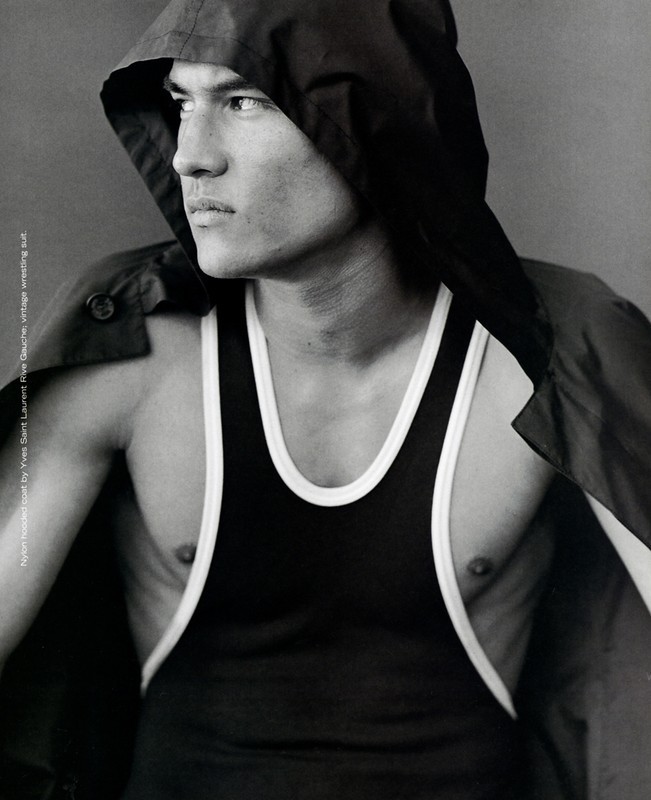 Monochrome portrait of a muscular adult male wearing a black top and hooded jacket, gazing to the side against a solid background.