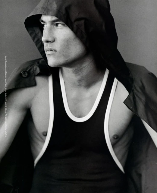 Monochrome portrait of a muscular adult male wearing a black top and hooded jacket, gazing to the side against a solid background.