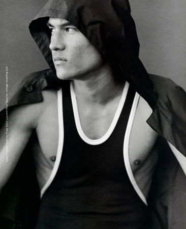 Man wearing a black hooded coat over a black tank top with white trim, looking to the side against a neutral background.