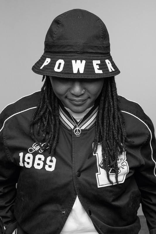 Monochrome portrait of a woman wearing a bucket hat with "POWER" on it and a varsity jacket, looking downward with a neutral expression.