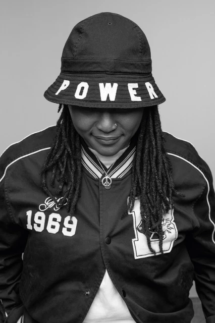 Monochrome portrait of a woman wearing a bucket hat with "POWER" on it and a varsity jacket, looking downward with a neutral expression.