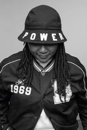 Monochrome portrait of a woman wearing a bucket hat with "POWER" on it and a varsity jacket, looking downward with a neutral expression.
