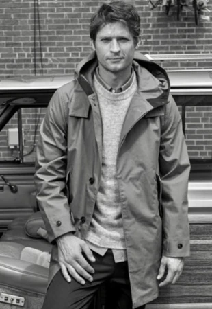 Man wearing hooded parka coat and knit sweater standing outdoors with a car and brick wall background
