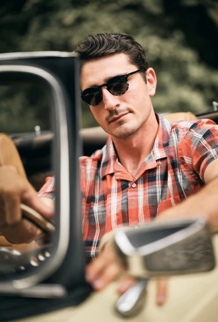 Portrait of a man in sunglasses and a red plaid shirt in a vintage car against a blurred green outdoor backdrop.