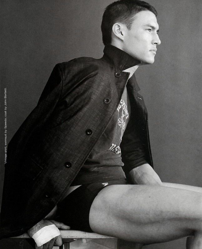 Monochrome portrait of a man in a coat and Speedo swimsuit sitting on a chair, looking to the side, showcasing a fashion editorial.