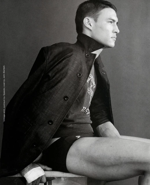 Monochrome portrait of a man in a coat and Speedo swimsuit sitting on a chair, looking to the side, showcasing a fashion editorial.