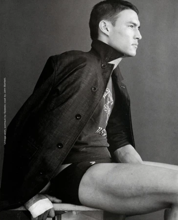 Monochrome portrait of a man in a coat and Speedo swimsuit sitting on a chair, looking to the side, showcasing a fashion editorial.