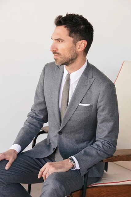 Portrait of an adult man in a gray suit and tie sitting on a wooden chair in a studio with a neutral background, looking off to the side.