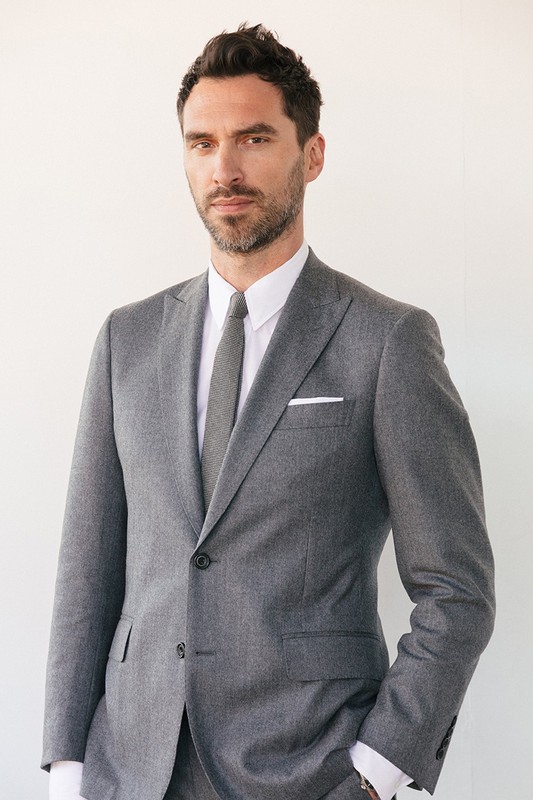 Headshot of an adult male wearing a grey suit with a white pocket square against a white background. Portrait orientation.
