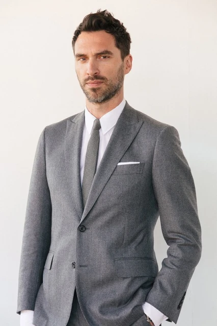 Headshot of an adult male wearing a grey suit with a white pocket square against a white background. Portrait orientation.