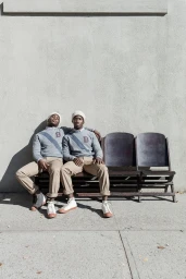 Two black men seated together on an outdoor bench wearing matching grey sweaters and khaki trousers.