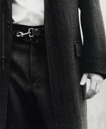 Close-up of a person wearing a grey tweed coat over trousers and a white shirt styled with a black leather belt.