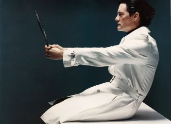 Man in white trench coat holding hand mirror in profile view against dark background