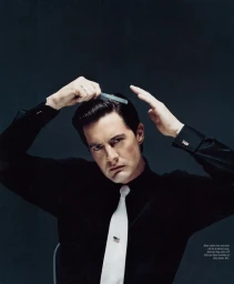 Man with dark hair combing his hair, wearing a black shirt and white tie with a tie clip against a dark textured background
