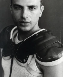 Close-up portrait of a man wearing a white tank top and dark protective athletic shoulder pads against a dark background.