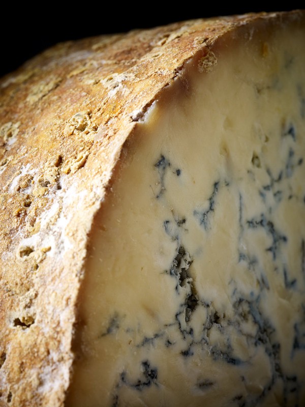 Close-up photograph of a wedge of blue cheese, showcasing its creamy texture, blue veins, and natural rind in a dark, shadowed setting.