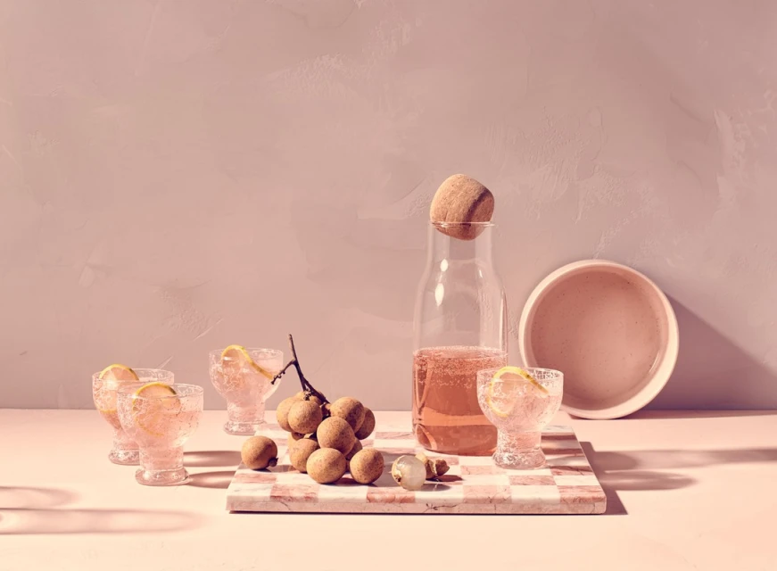 Still life composition featuring glasses of pink cocktails, a carafe with a stone stopper, fruit, and a checkered tray on a pink surface.