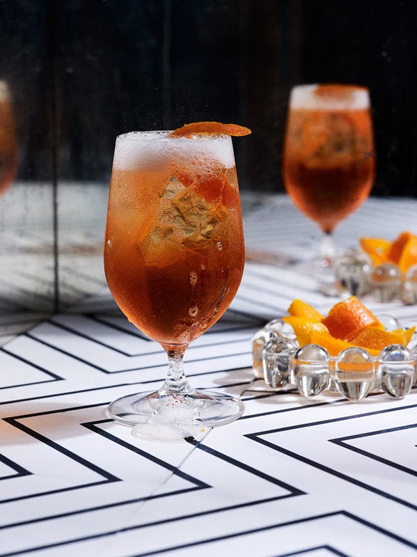 Two stemmed glasses filled with orange cocktails and topped with orange peels sit on a geometric surface in a dark, indoor setting.