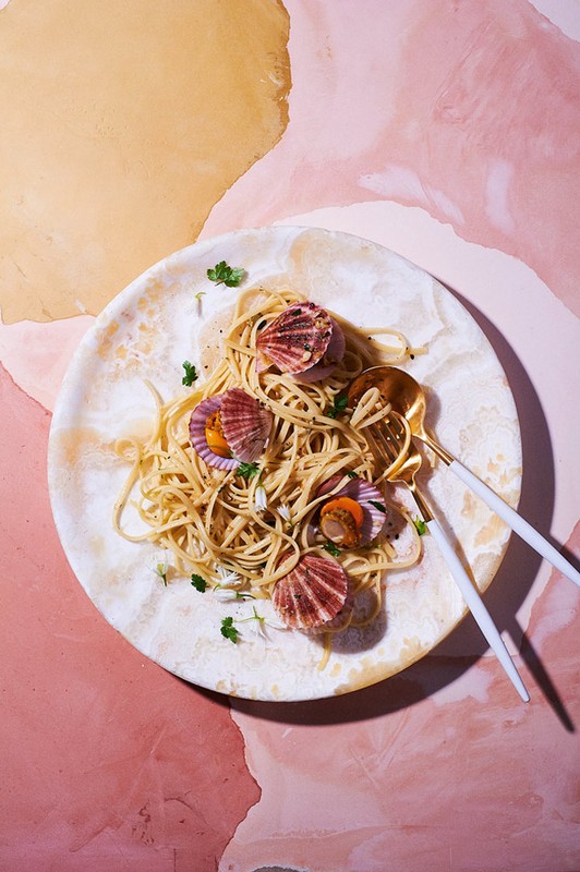 Overhead view of scallop pasta dish presented on a patterned plate with gold serving utensils, set against a pink and tan background.