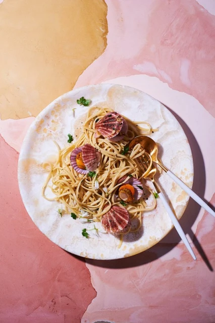 Overhead view of scallop pasta dish presented on a patterned plate with gold serving utensils, set against a pink and tan background.