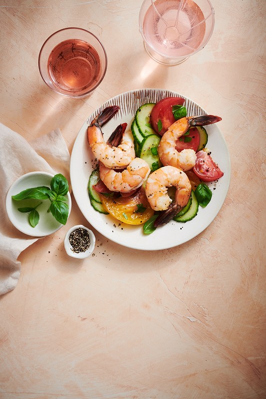 Overhead shot of a plate of shrimp salad with tomatoes, cucumbers, basil, and rose wine on a light pink textured surface.