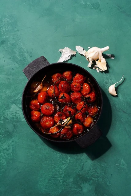 Overhead shot of roasted cherry tomatoes in a black pan, with garlic cloves on a textured teal background, bathed in natural light.