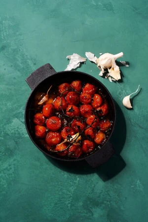 Overhead shot of roasted cherry tomatoes in a black pan, with garlic cloves on a textured teal background, bathed in natural light.