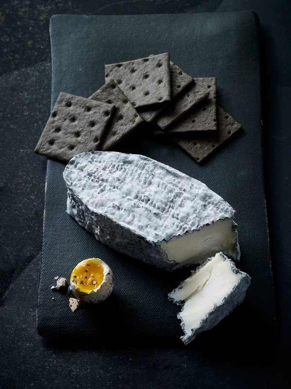 Overhead shot of a slate board with cheese, crackers, and an egg. The composition features cool tones and contrasts textures.
