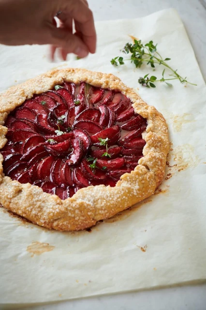 Overhead shot of a rustic plum galette on parchment paper, garnished with fresh thyme, and a hand adding greens.