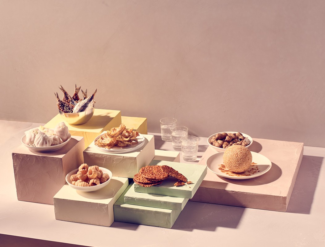 Still life of diverse Asian snacks on colorful geometric platforms, with clear glasses of water and a light pink background.
