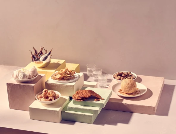Still life of diverse Asian snacks on colorful geometric platforms, with clear glasses of water and a light pink background.