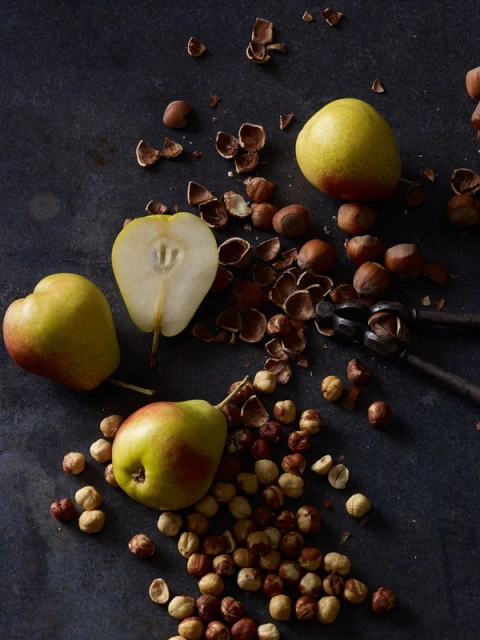 Overhead shot of pears, hazelnuts, and a nutcracker on a dark surface, showcasing rustic food styling and natural light.